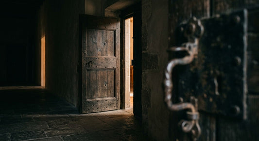 Une porte ancienne en bois entrouverte, lumière chaude filtrant dans un couloir de pierre sombre